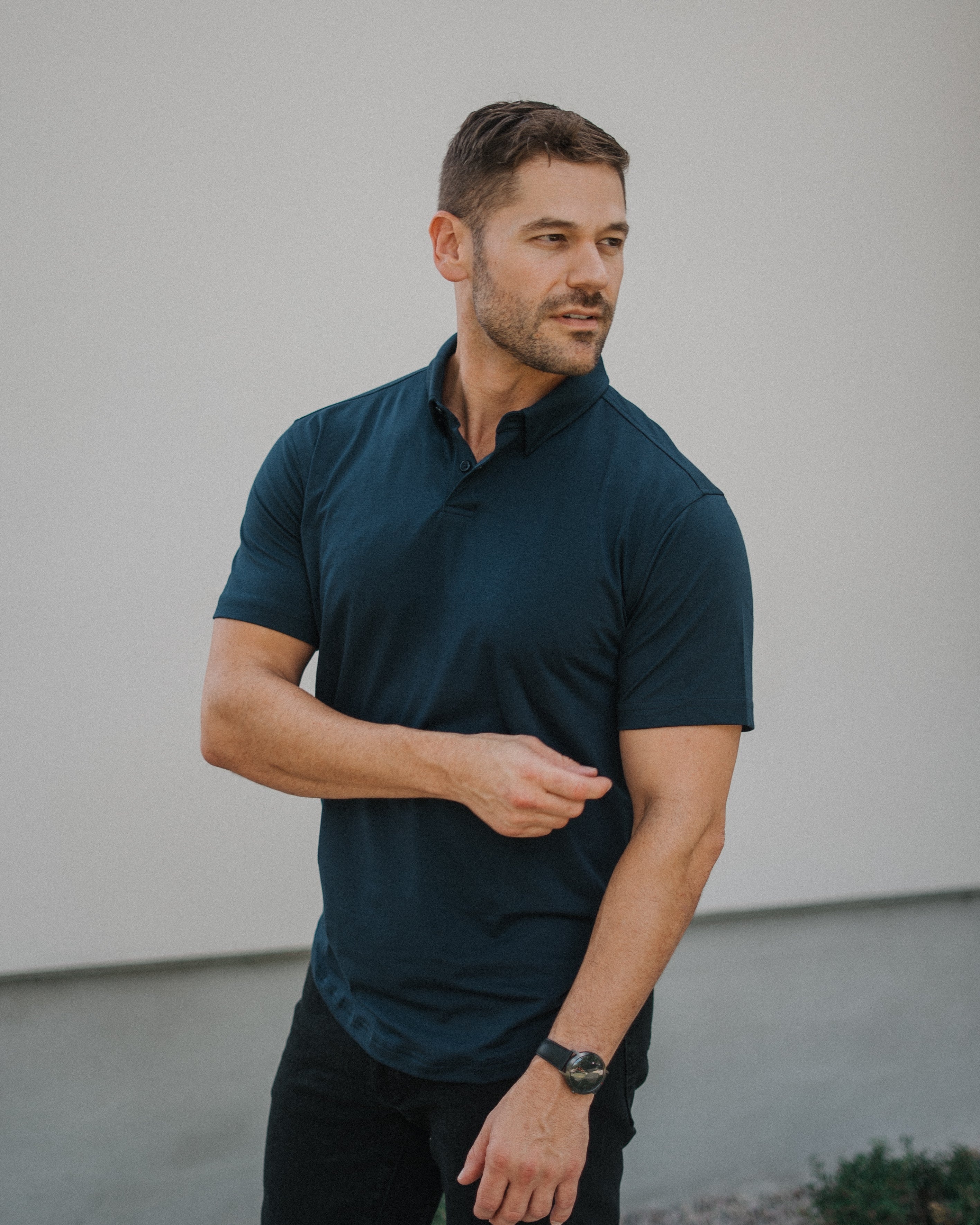 A man wearing a dark navy blue short-sleeve polo shirt and black pants, standing outdoors against a light gray wall.