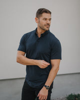 A man wearing a dark navy blue short-sleeve polo shirt and black pants, standing outdoors against a light gray wall.