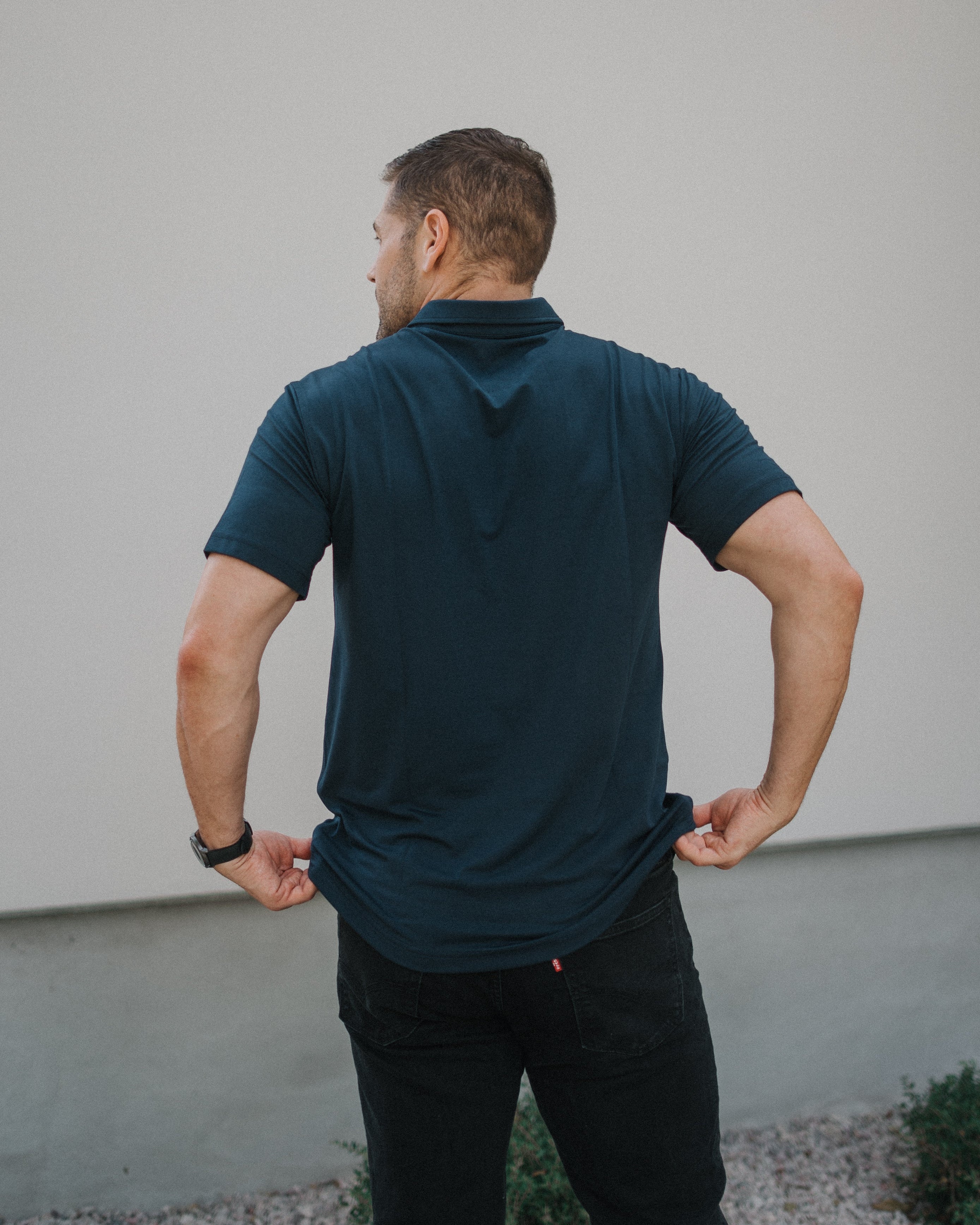 A man viewed from behind wearing a dark teal short-sleeve polo shirt and black pants, standing outdoors against a light gray wall.