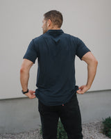 A man viewed from behind wearing a dark teal short-sleeve polo shirt and black pants, standing outdoors against a light gray wall.