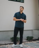 A man wearing a dark navy blue short-sleeve polo shirt, black pants, and white sneakers stands outdoors in front of a beige stucco wall.