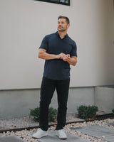 A man wearing a dark navy blue short-sleeve polo shirt, black pants, and white sneakers stands outdoors in front of a beige stucco wall.