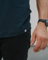 Close-up of a person wearing a dark navy blue t-shirt and black jeans, with a small white logo on the bottom hem of the shirt. The person is also wearing a black watch.