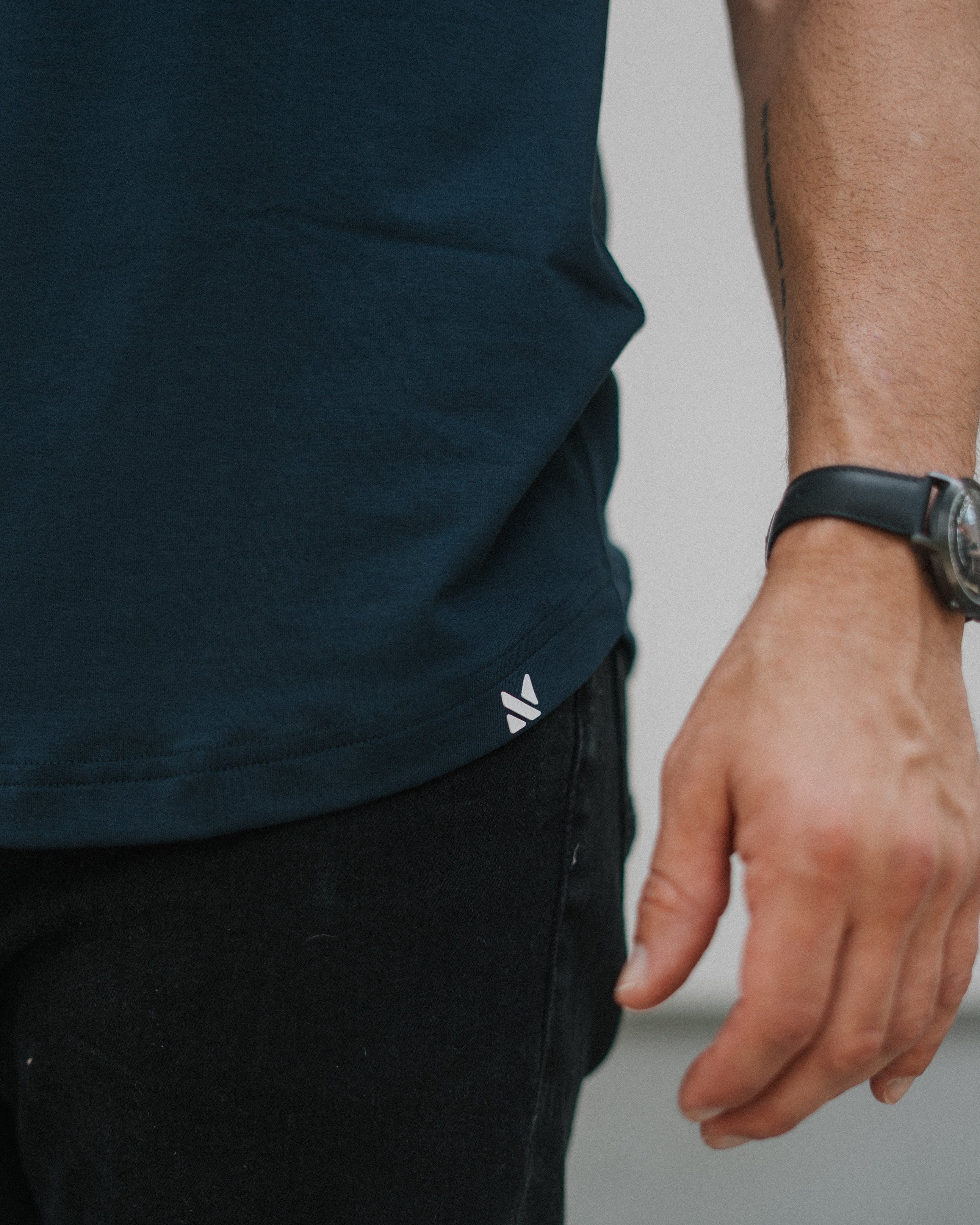 Close-up of a person wearing a dark navy blue t-shirt and black jeans, with a small white logo on the bottom hem of the shirt. The person is also wearing a black watch.