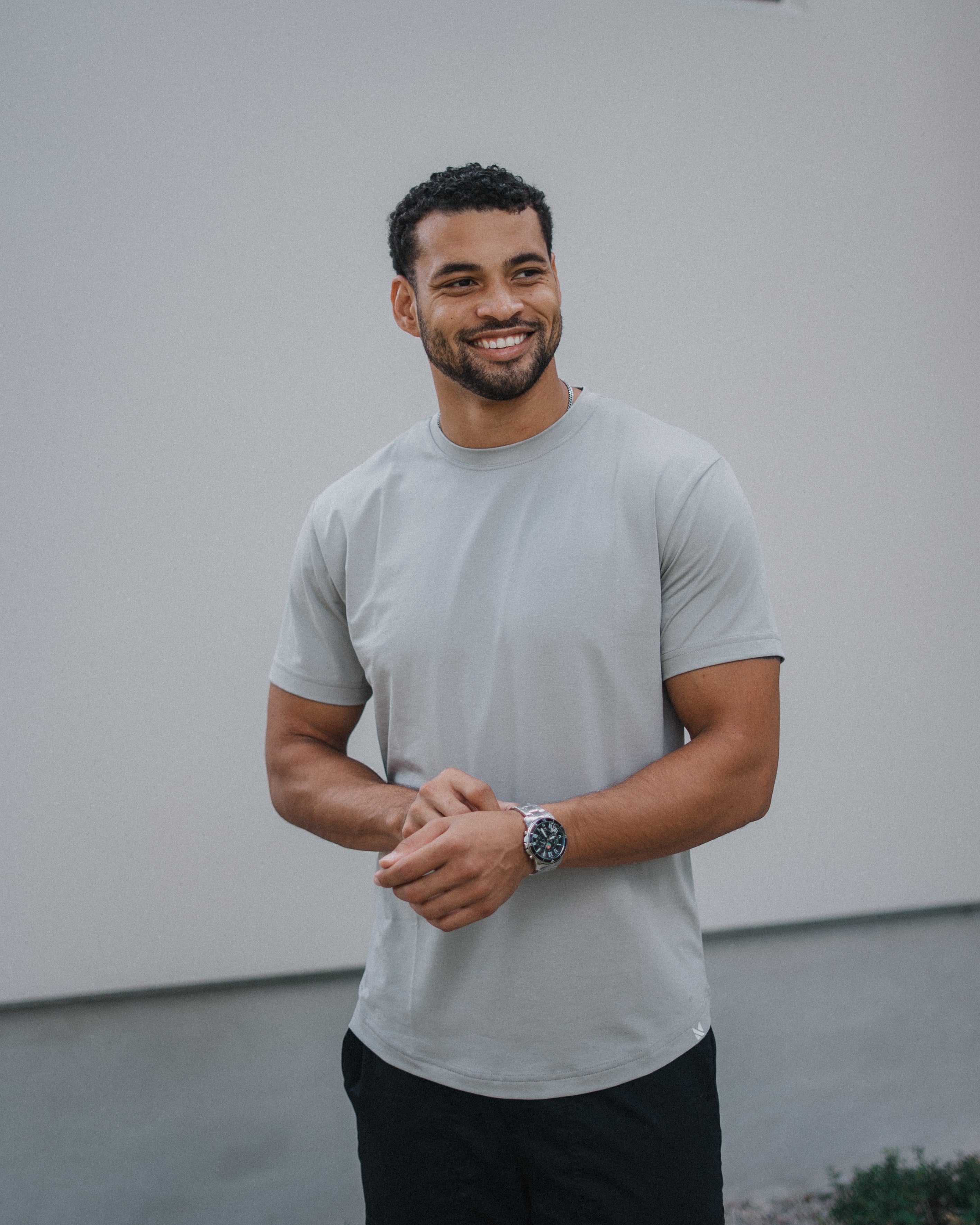 A smiling man wearing a light gray crewneck t-shirt and black pants, standing outdoors against a white wall.