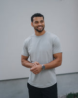 A smiling man wearing a light gray crewneck t-shirt and black pants, standing outdoors against a white wall.