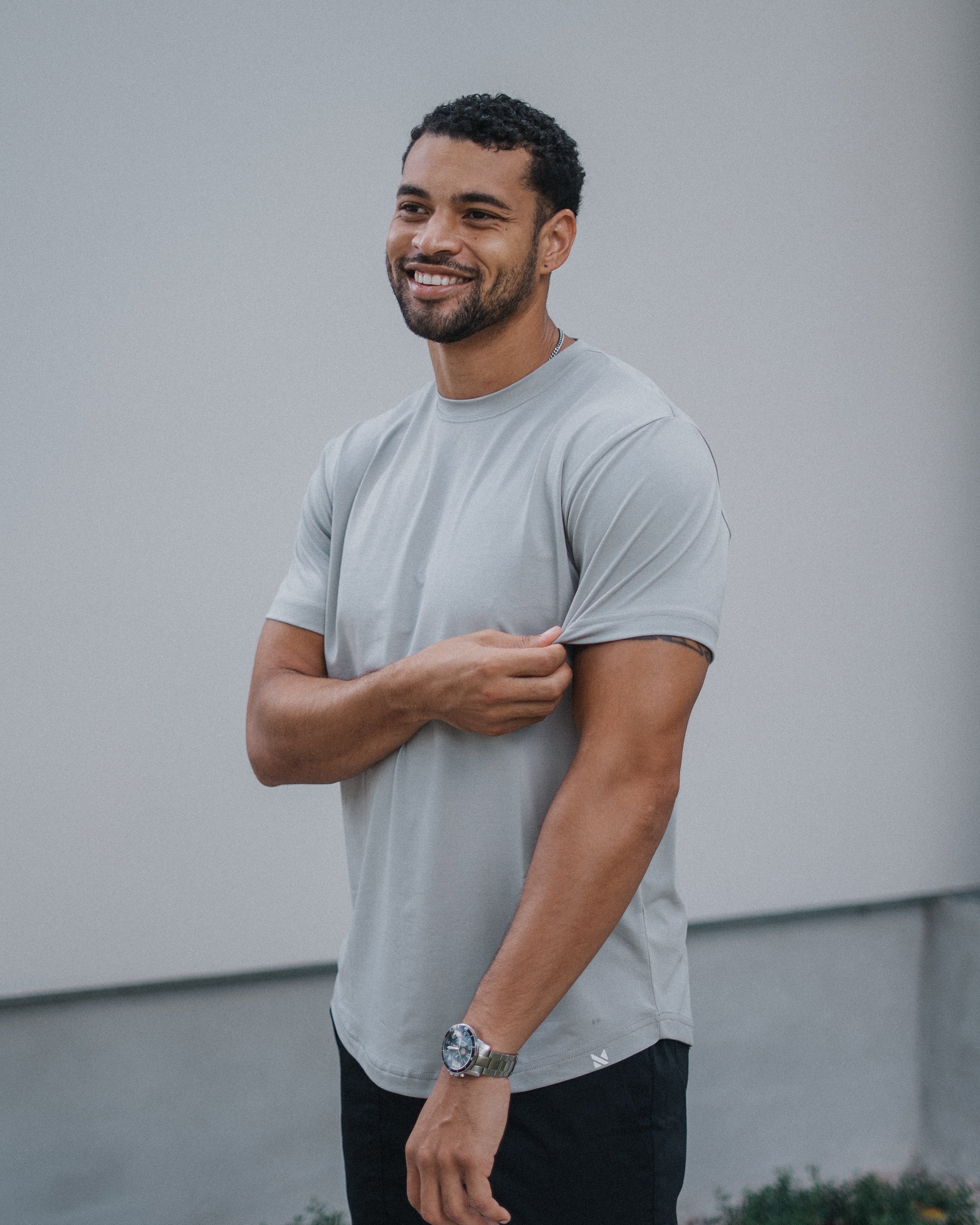 A smiling man wearing a light gray crewneck t-shirt and black pants, standing against a light gray wall.