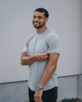 A smiling man wearing a light gray crewneck t-shirt and black pants, standing against a light gray wall.