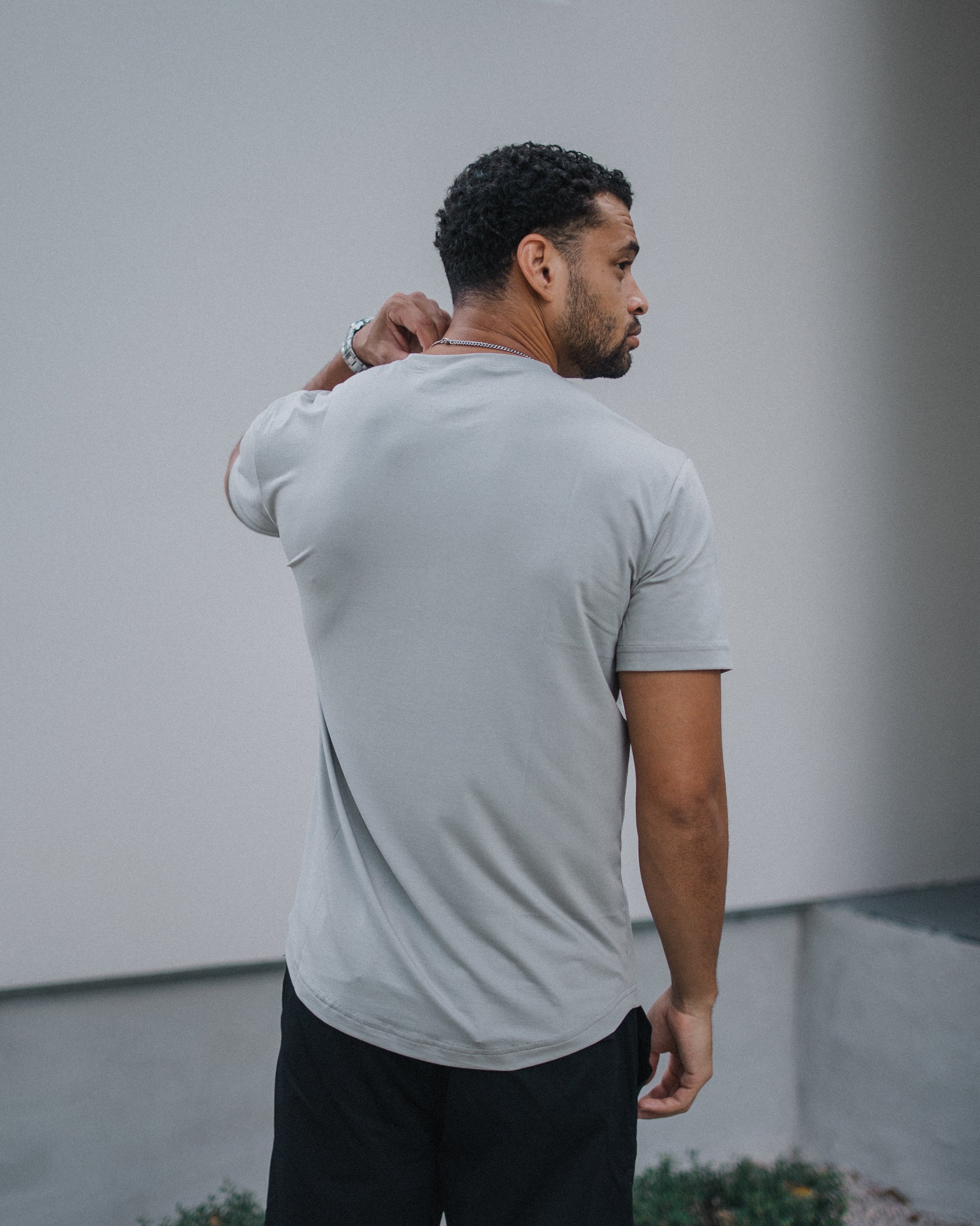 A man wearing a light gray short-sleeve t-shirt and black pants, standing outdoors against a modern light gray wall.