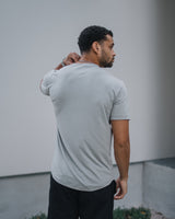 A man wearing a light gray short-sleeve t-shirt and black pants, standing outdoors against a modern light gray wall.