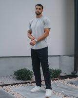 A man wearing a light gray crewneck t-shirt, black cargo pants, and white sneakers, standing outdoors in front of a modern house wall.