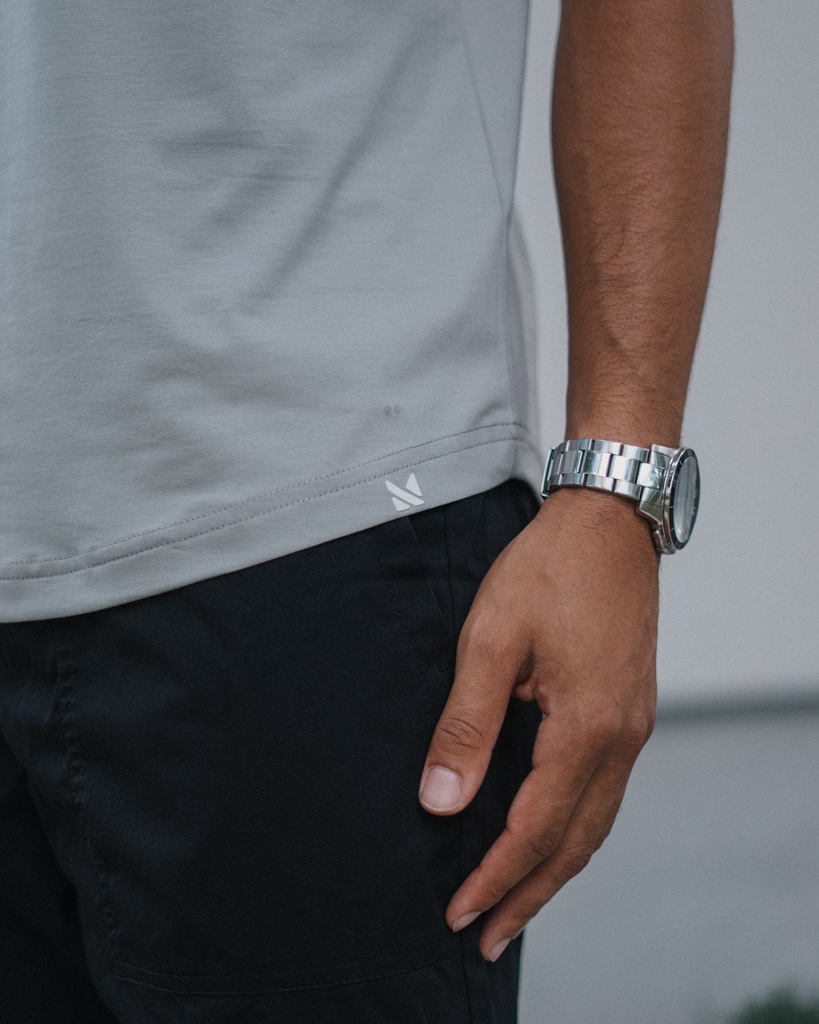 Close-up of a person wearing a light gray t-shirt, black pants, and a silver metal link watch on their wrist.