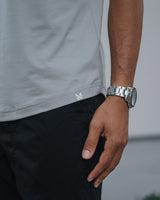 Close-up of a person wearing a light gray t-shirt, black pants, and a silver metal link watch on their wrist.