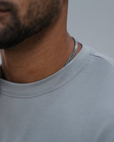 Close-up of a man's neck and lower face wearing a silver chain necklace and a light gray crew-neck t-shirt.