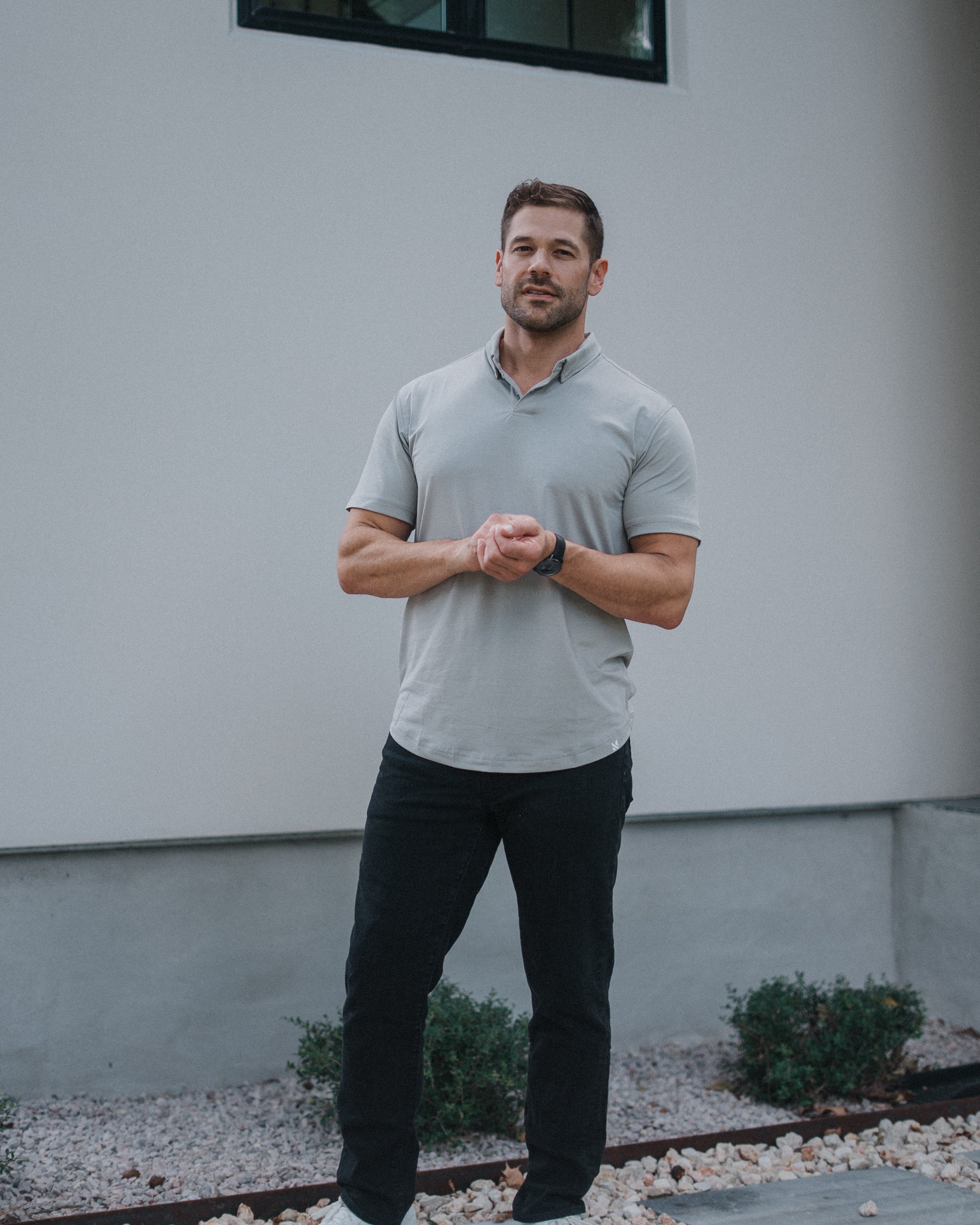 A man wearing a light gray short-sleeve polo shirt, black pants, and white sneakers stands outdoors in front of a modern gray stucco building.