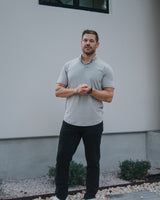 A man wearing a light gray short-sleeve polo shirt, black pants, and white sneakers stands outdoors in front of a modern gray stucco building.