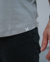 Close-up of a person wearing a light gray t-shirt and black jeans, showing a small white geometric logo on the bottom hem of the shirt.