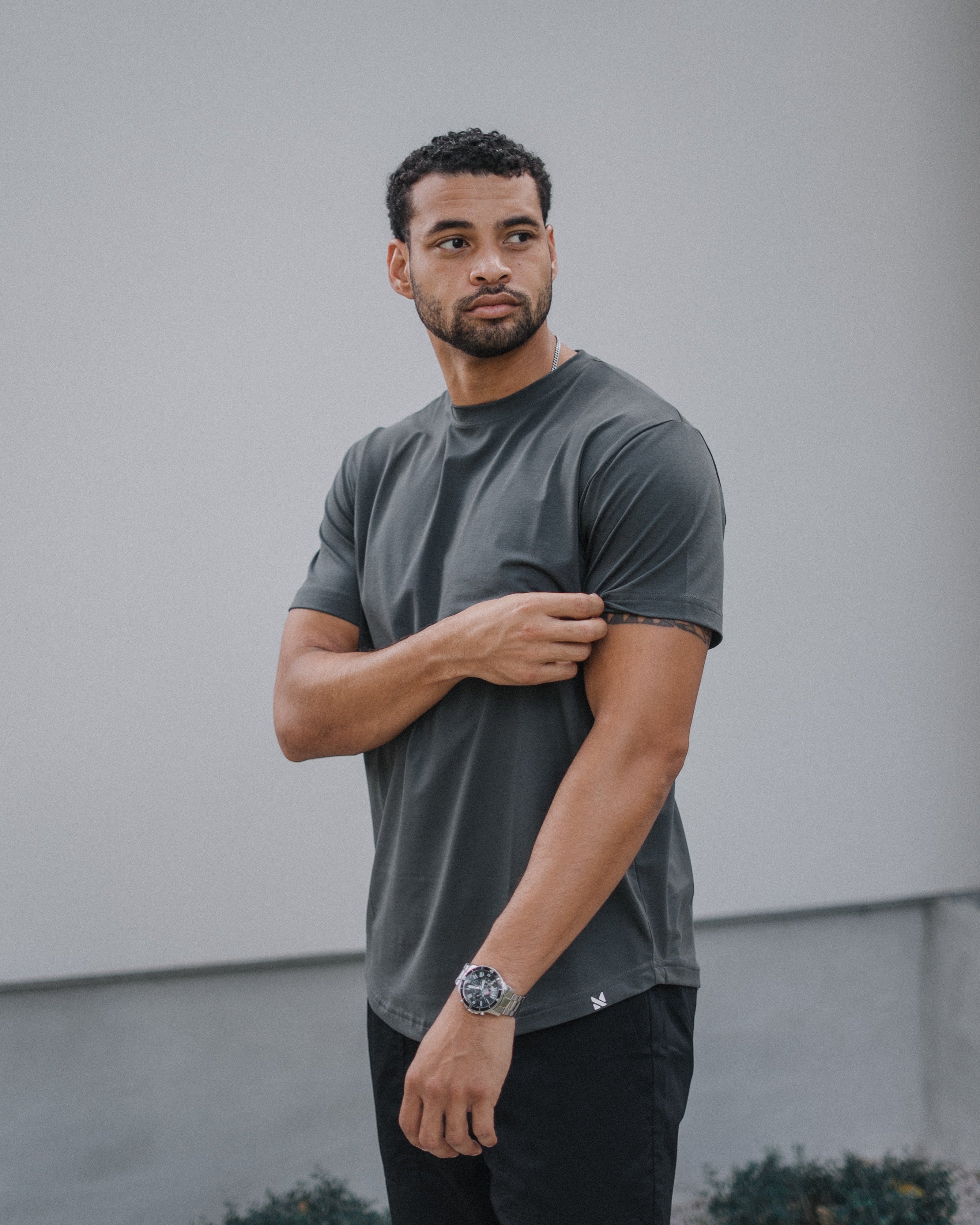 A man wearing a dark gray crewneck t-shirt and black pants, standing outdoors against a light gray wall.