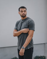 A man wearing a dark gray crewneck t-shirt and black pants, standing outdoors against a light gray wall.