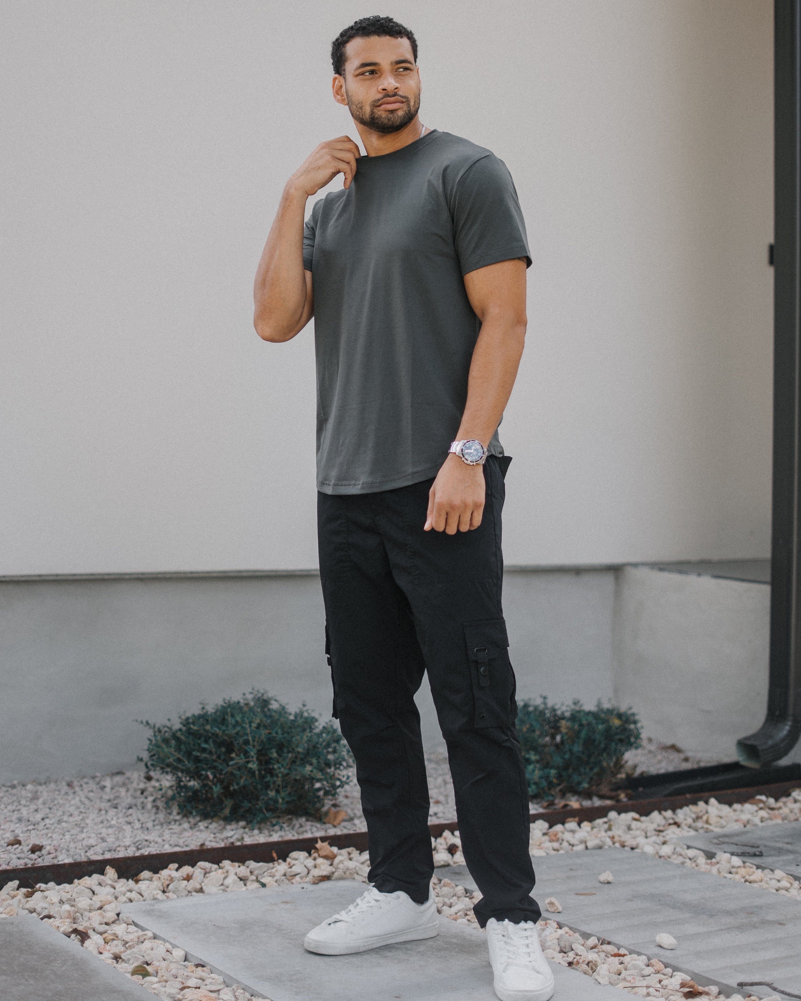 A man wearing a dark gray crewneck t-shirt, black cargo pants, and white sneakers, standing outdoors in front of a modern house wall.