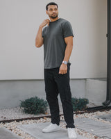 A man wearing a dark gray crewneck t-shirt, black cargo pants, and white sneakers, standing outdoors in front of a modern house wall.