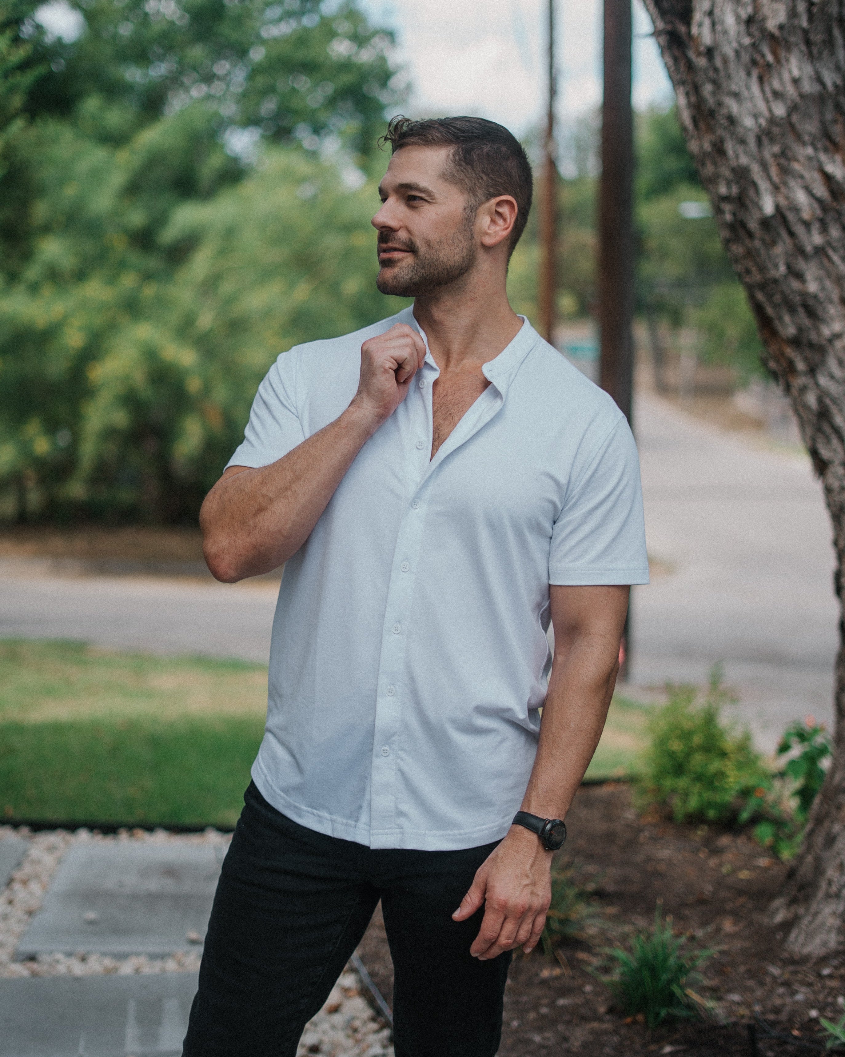 A man wearing a light blue short-sleeve button-down shirt and black pants, standing outdoors next to a tree.
