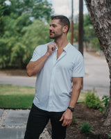 A man wearing a light blue short-sleeve button-down shirt and black pants, standing outdoors next to a tree.