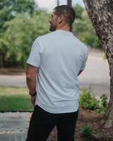 A man viewed from behind wearing a light gray short-sleeve t-shirt and black pants, standing outdoors next to a large tree trunk.
