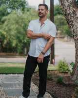 A man wearing a light blue short-sleeve button-down shirt, black pants, and white sneakers stands on a stone path outdoors next to a large tree trunk.