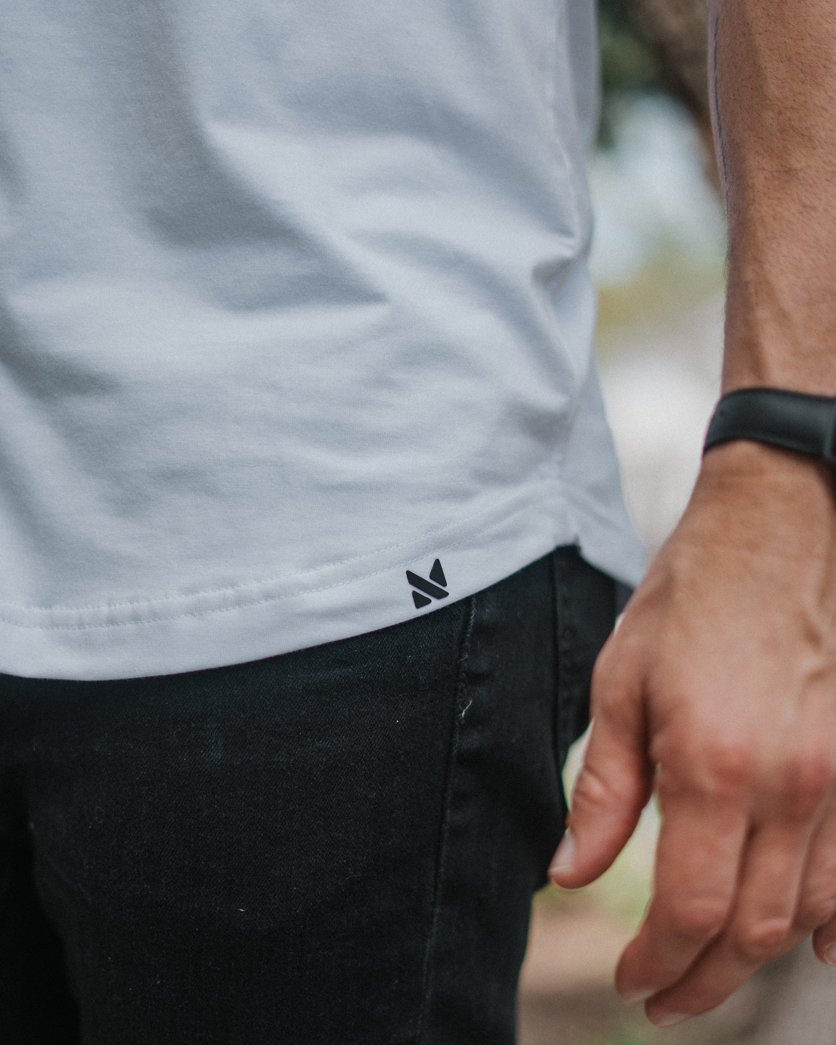 Close-up of a person wearing a white t-shirt with a small black geometric logo near the hem, paired with black jeans and a black smartwatch.