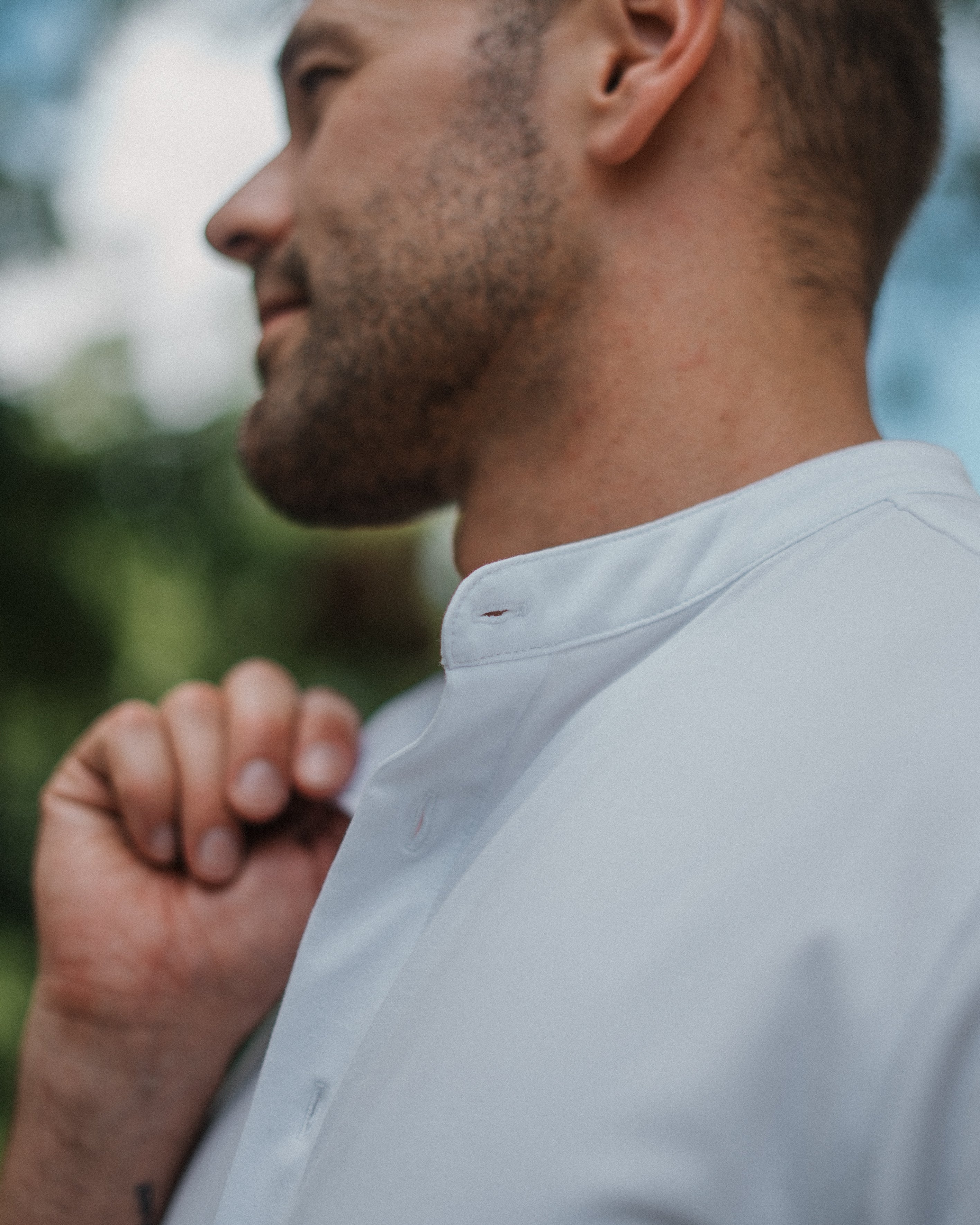 A close-up side profile of a man wearing a light gray or off-white button-up shirt, with his hand near his collar, set against a soft, blurred outdoor background.