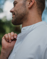 A close-up side profile of a man wearing a light gray or off-white button-up shirt, with his hand near his collar, set against a soft, blurred outdoor background.