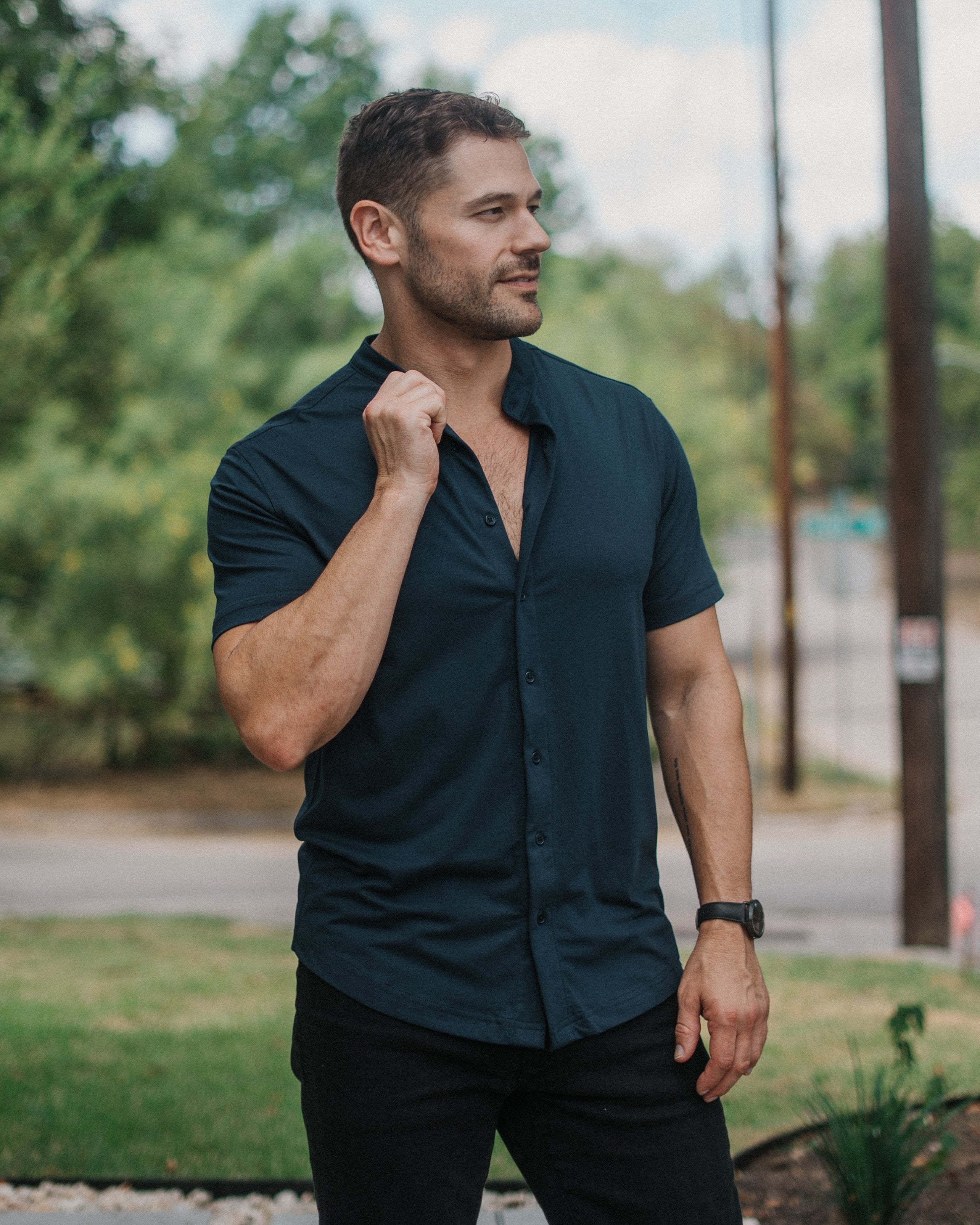 A man wearing a dark navy blue short-sleeve button-down shirt and black pants, standing outdoors in a grassy area with trees and utility poles in the background.