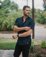 A man wearing a dark navy short-sleeve button-up shirt and black pants, standing outdoors in a residential area with trees, a utility pole, and a paved walkway.