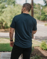 A man viewed from behind wearing a dark navy blue crewneck t-shirt and black pants, standing outdoors on a paved path with trees and a street in the background.