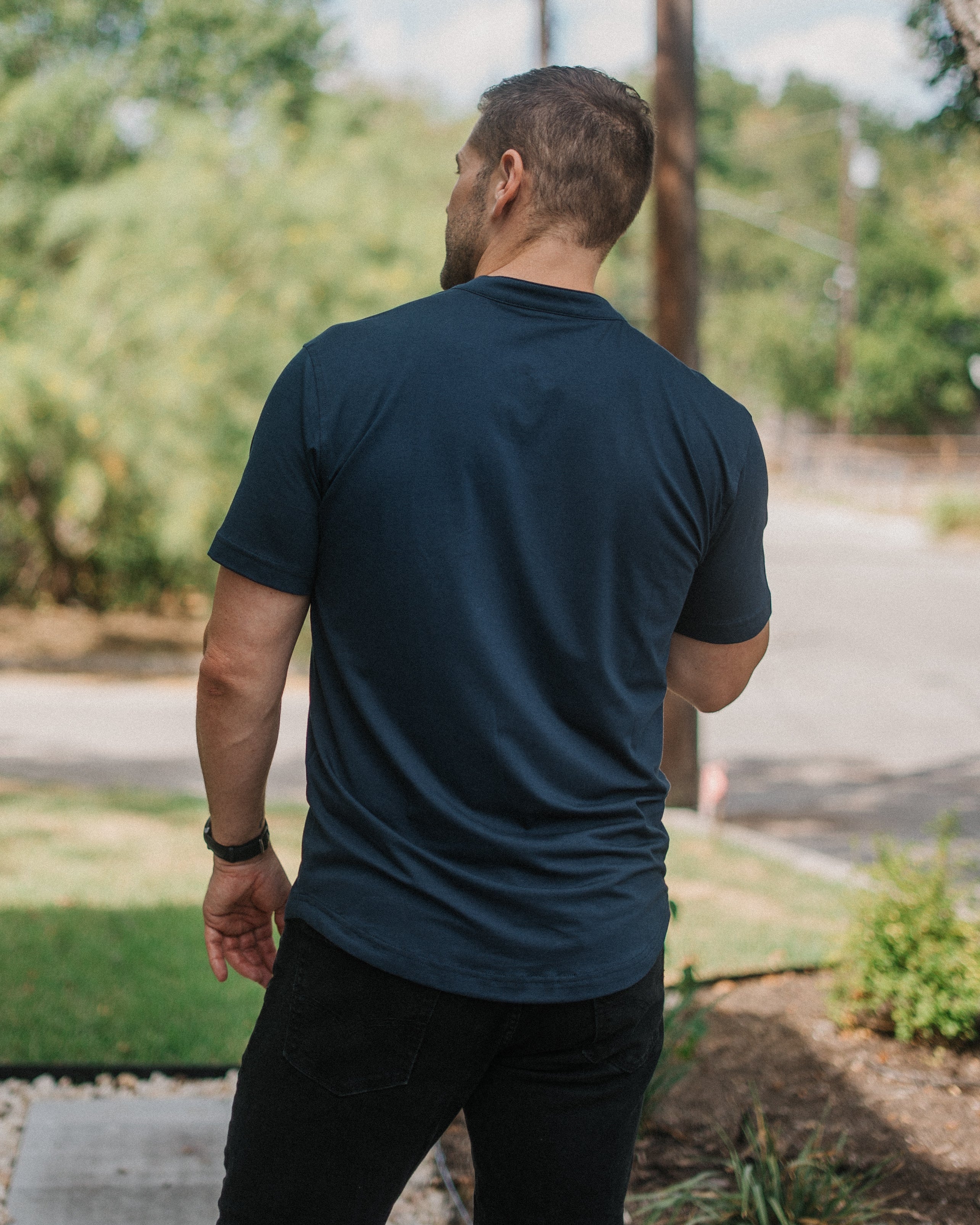 A man viewed from behind wearing a dark navy blue crewneck t-shirt and black pants, standing outdoors on a paved path with trees and a street in the background.