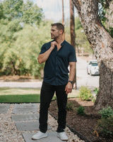 A man wearing a dark navy blue short-sleeve button-down shirt, black pants, and white sneakers stands outdoors on a stone path next to a large tree trunk.
