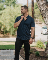 A man wearing a dark navy blue short-sleeve button-down shirt, black pants, and white sneakers stands outdoors on a stone path next to a large tree trunk.