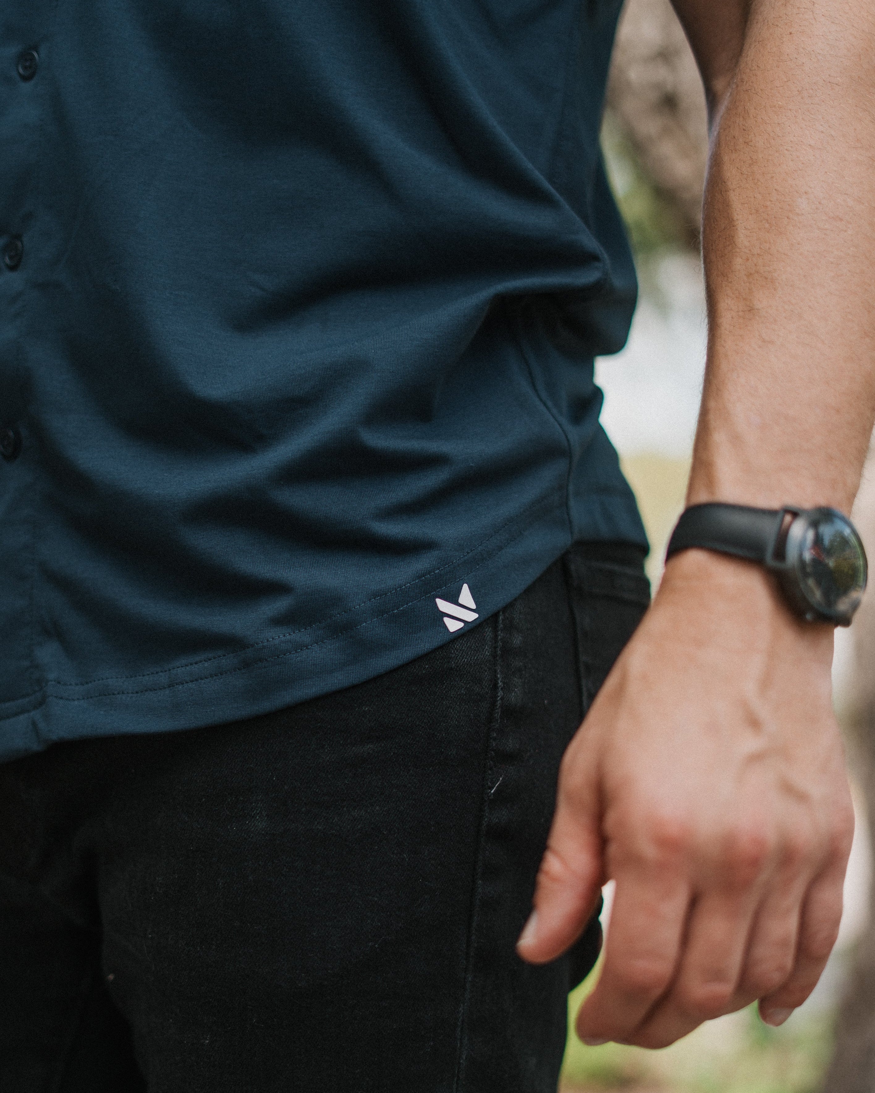 Close-up of a person wearing a dark navy button-up shirt and black pants, with a small white logo near the hem and a black wristwatch visible on the arm.