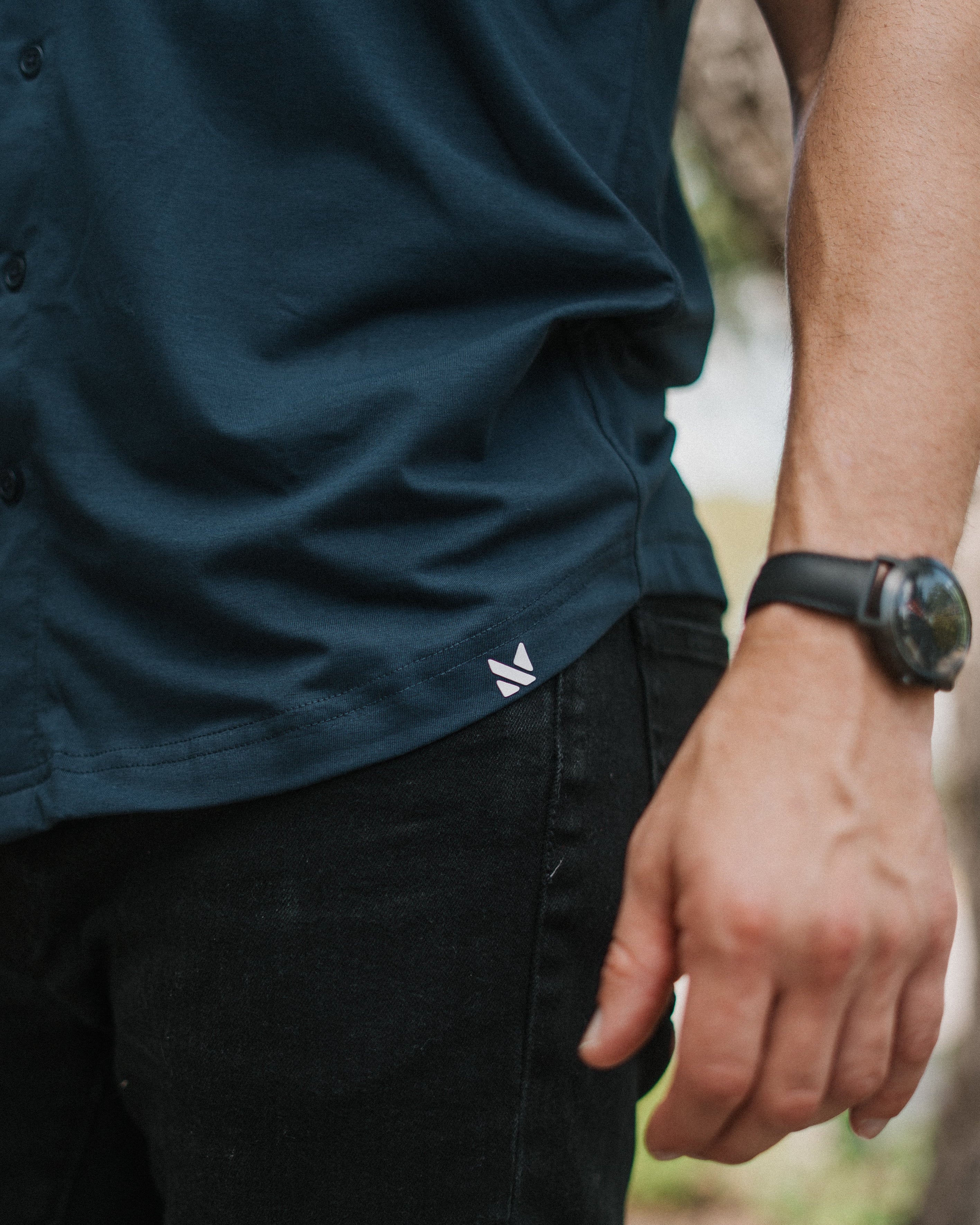 Close-up of a person wearing a dark navy button-up shirt and black pants, with a small white logo near the hem and a black wristwatch visible on the arm.