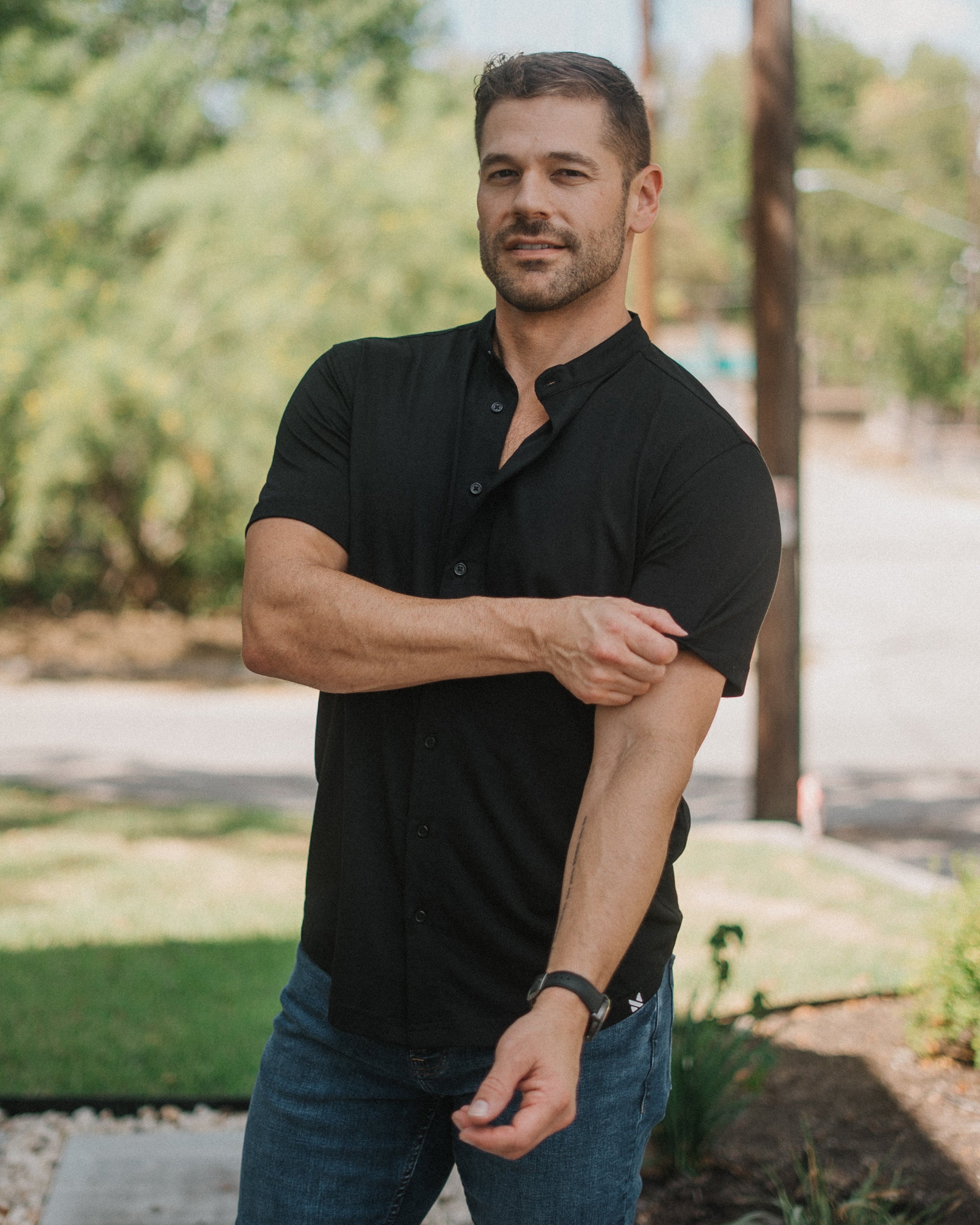 A man wearing a black short-sleeve button-down shirt and blue jeans standing outdoors in a sunny, grassy area with trees and a utility pole in the background.