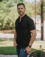 A man wearing a black short-sleeve polo shirt and blue jeans standing outdoors in a sunny residential setting with trees, a utility pole, and a stone walkway.