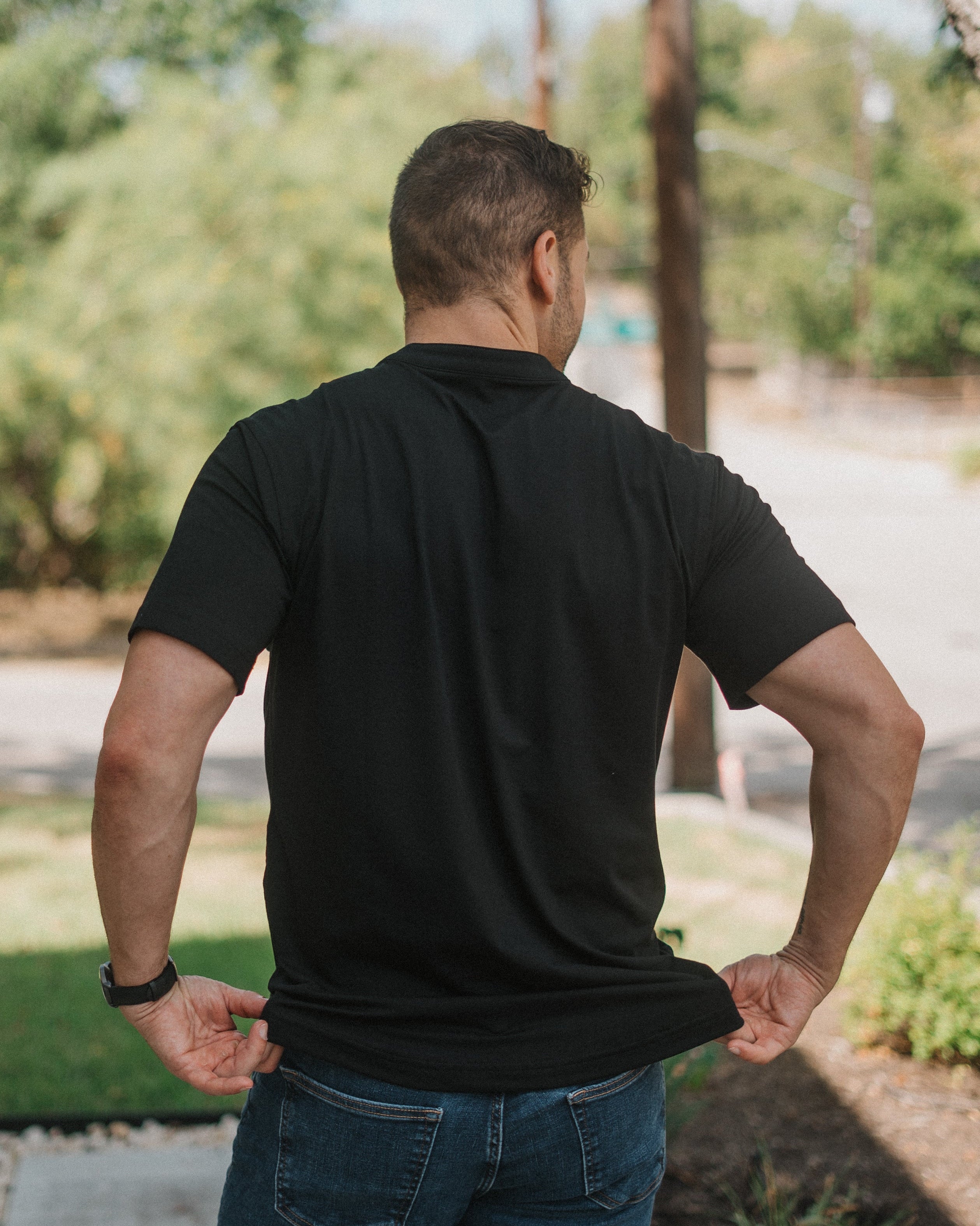 A man viewed from behind wearing a plain black short-sleeve polo shirt and blue jeans, standing outdoors on a sunny day with trees and a path in the background.