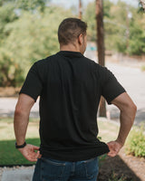 A man viewed from behind wearing a plain black short-sleeve polo shirt and blue jeans, standing outdoors on a sunny day with trees and a path in the background.