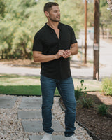A man wearing a black short-sleeve button-up shirt, blue jeans, and white sneakers stands on a stone path outdoors near a tree and gravel ground.