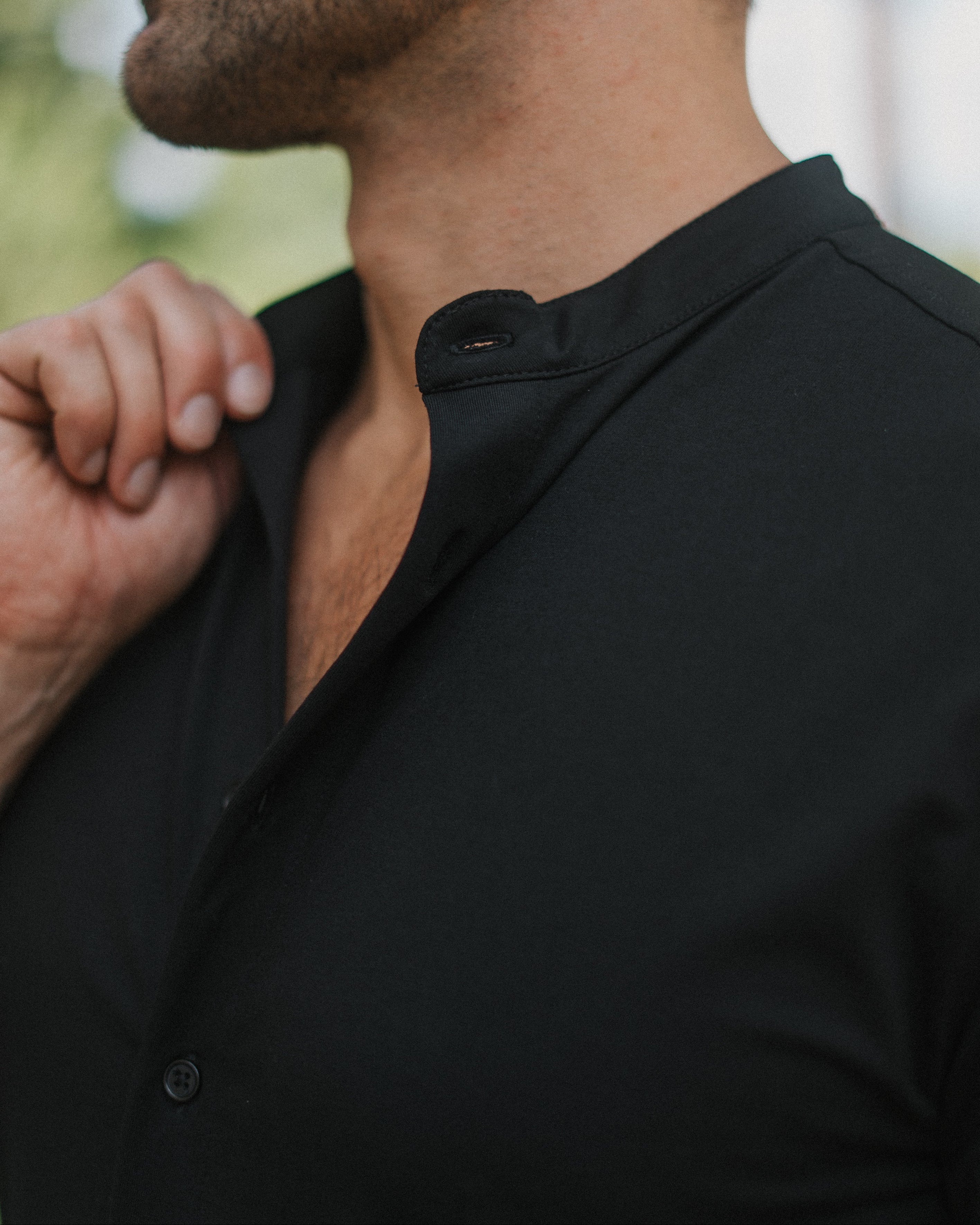 Close-up of a man wearing a black long-sleeve button-up shirt with a mandarin collar, shown from the side in an outdoor setting.
