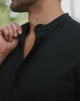 Close-up of a man wearing a black long-sleeve button-up shirt with a mandarin collar, shown from the side in an outdoor setting.