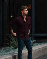 Man in a burgundy shirt and jeans standing outdoors with a dark background