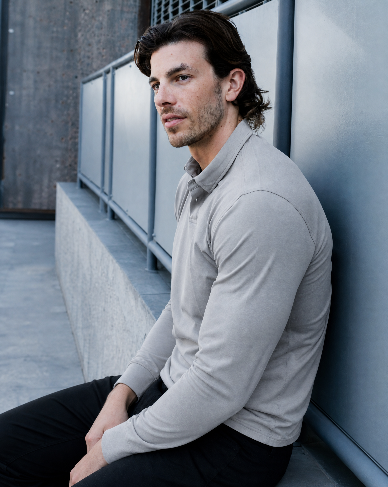 Man sitting against a wall wearing a light gray sweater and dark pants.