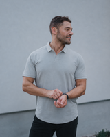 A man wearing a light gray short-sleeve polo shirt and black pants, standing outdoors against a gray wall.